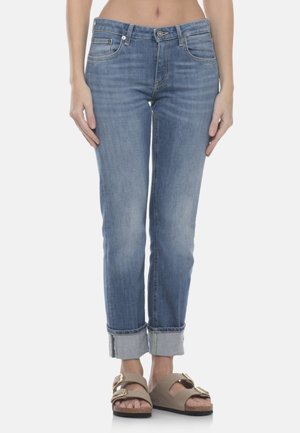 Donna che indossa jeans in denim azzurro con risvolti e sandali beige con due fibbie d'oro, in piedi davanti a uno sfondo semplice.