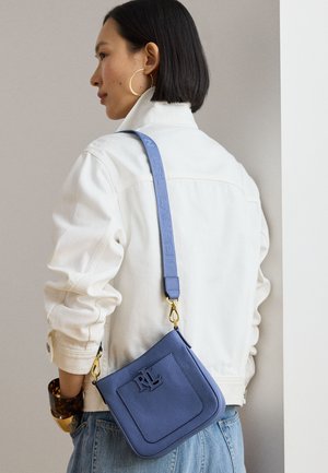 Nainen valkoisessa takissa ja sinisissä farkuissa kantaa sinistä nahkaista olkalaukkua, jossa on kultaiset metalliosat ja säädettävä kangashihna.