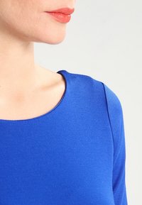 Haut à manches longues bleu avec une texture lisse, encolure ronde et design ajusté, mettant en valeur une couleur riche et des détails d'épaule sans couture.