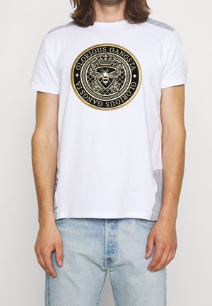Mand iført hvid t-shirt med cirkulært sort og gyldent emblem med en bi og teksten "Glorious Gangsta" i midten.