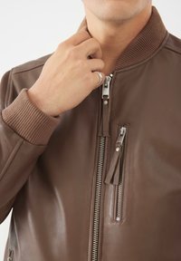 Giacca bomber in pelle marrone con polsini e colletto a costine, con cerniere argentate sul davanti e nelle tasche laterali, texture liscia.