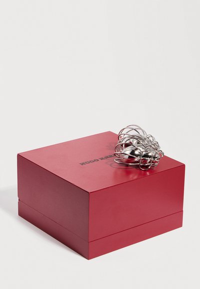 Κόκκινο κουτί Hugo Kretschmer σε σχήμα τετραγώνου με ασημένιο αφηρημένο μεταλλικό γλυπτό που ακουμπά πάνω του, σε λευκό φόντο.