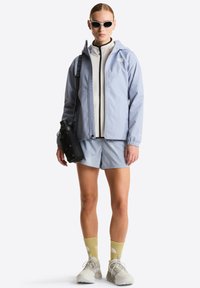 Γυναίκα που φοράει ανοιχτό μπλε μπουφάν και σορτς, μπεζ φλις με φερμουάρ, κίτρινες κάλτσες, άσπρα αθλητικά παπούτσια, άσπρα γυαλιά ηλίου, κρατώντας μια μαύρη τσάντα για παγούρι.