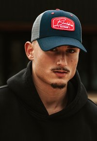 Tengerészkék sapka hálós hátsó résszel és piros hímzett logóval, amelyet egy fekete kapucnis pulóvert viselő személy hord.