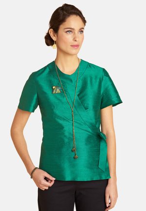 Frau trägt eine glänzende grüne Wickelbluse mit kurzen Ärmeln und Brosche, lange goldene Halskette, schwarze Hose und ein Armband, steht vor einem weißen Hintergrund.