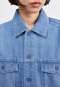 Denim jacket in light blue, featuring a collared neckline, snap buttons, two chest pockets, and a textured, slightly faded finish.