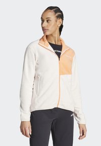 Chaqueta de forro polar ligera en color crema, con cremallera naranja y panel en el pecho, tela texturizada y dos bolsillos laterales. Cremallera delantera oculta.