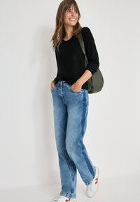 Jeans in denim azzurro chiaro con una vestibilità ampia, abbinati a un top nero a maniche lunghe. Sneakers bianche con dettagli rossi. Uno zaino verde oliva è appoggiato su una spalla.