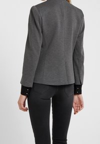 Blazer gris ajusté avec un col haut, doté de poignets à boutons noirs et d'un tissu texturé. Porté sur un pantalon sombre. Vue de dos.