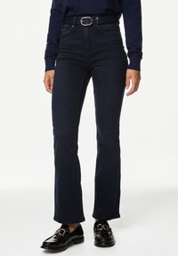 Mujer vestida con jeans ajustados de color azul oscuro, cinturón negro, blusa de manga larga azul marino y mocasines negros con hebillas doradas, de pie sobre un fondo blanco.