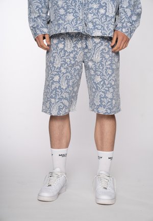 Mann trägt blaue Shorts mit Paisley-Muster und passendes langärmeliges Hemd, weiße Socken mit schwarzem Text und weiße Turnschuhe, steht.