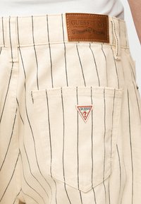 Μπεζ παντελόνι με ρίγες, με πίσω τσέπη που διαθέτει τριγωνικό λογότυπο GUESS και καφέ δερμάτινη ετικέτα της μάρκας GUESS JEANS στη ζώνη μέσης.