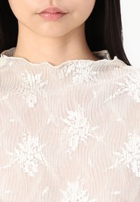 Top de renda em creme suave com um padrão floral, apresentando uma textura ondulada e um decote em forma de escalope. O tecido é leve e semi-transparente.