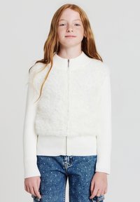 Felpa bianca con zip e un pannello anteriore fluffy, maniche e bordo a coste, abbinata a jeans blu con pois bianchi.