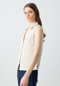 Canottiera in lino senza maniche di un leggero beige con colletto, scollo a V profondo e chiusura con bottoni, dotata di spacchi laterali per facilitare i movimenti.