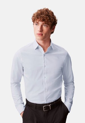 Joven con pelo rizado que lleva una camisa de botones azul claro y pantalones negros, con las manos en los bolsillos, mirando ligeramente hacia un lado.