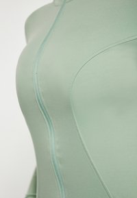 Indumento aderente verde chiaro realizzato in materiale liscio e elastico, dotato di chiusura a zip e cuciture curve per una silhouette slanciata.