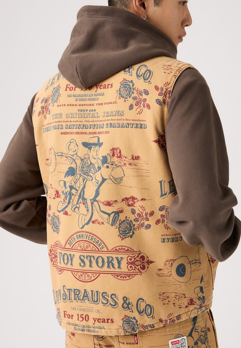 Tan denim vest med farverige trykte design, der indeholder figurer og tekst, parret med en brun hættetrøje.