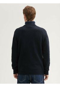 Marineblauwe trui met hoge kraag, geribde manchetten en zoom, een gestructureerd breisel en een eenvoudig ontwerp, gedragen over blauwe spijkerbroek.