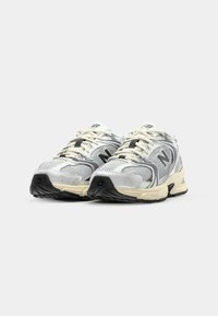 Scarpe da ginnastica grigie e bianche con pannelli in rete, suole in gomma e lacci. Presentano un logo nero prominente sul lato e un design strutturato.