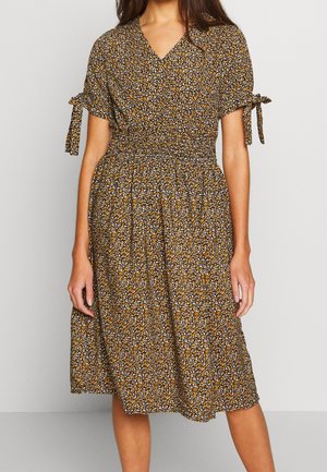 Kvinde iført en knælængde, kortærmet brun kjole med lille hvidt og orange blomsterprint og bindedetaljer på ærmerne.