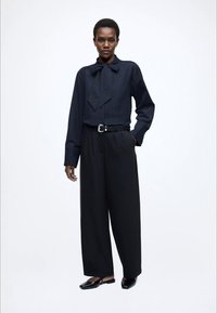 Tmavomodrá blúzka s textúrou a mašľou vpredu, skombinovaná s čiernymi širokými nohavicami s vysokým pásom a čiernymi plochými topánkami.