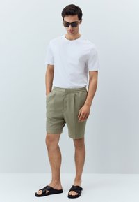 White short-sleeve t-shirt, olive green tailored shorts with pockets, and black slide sandals. Smooth fabric with a relaxed fit.
