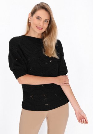 Pull en tricot noir avec un motif en vagues, manches courtes et un col large, porté sur un pantalon beige clair. Détails de tricot texturé visibles.