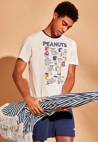 Jeune homme portant un t-shirt blanc Peanuts et un short bleu marine, tenant une couverture pliée à rayures bleues et blanches avec des franges.