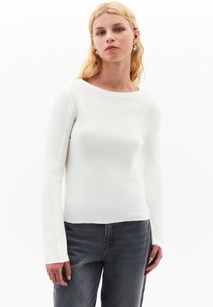 Frau mit blonden Haaren, die silberne Creolen, ein weißes, langärmliges eng anliegendes Oberteil und dunkelgraue Jeans vor einem weißen Hintergrund trägt.