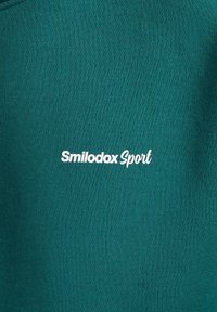 Turkusowy sweter z gładką fakturą z napisem "Smilodox Sport" w białym haftem na piersi.