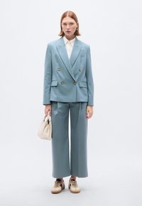 Σακάκι διπλής breasted σε φωτεινό γαλάζιο, παντελόνια με φαρδύ πόδι και εκρού πουκάμισο. Κρατά μια μικρή μπεζ τσάντα.