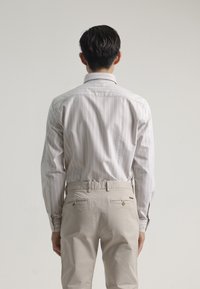 Camisa de manga larga a rayas en gris claro con rayas blancas. Pantalones beiges con costuras y detalles de botones. Vista desde la espalda.