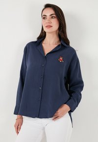 Navy blue button-up shirt with long sleeves, featuring an embroidered bear design on the left chest. Made of lightweight fabric, relaxed fit.