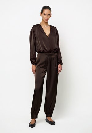 Vrouw met donker haar in een strakke knot, die een donkerbruine satijnen blouse draagt met diepe V-hals en bijpassende satijnen broek met zwarte platte schoenen.