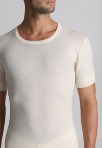 Uomo che indossa una maglietta a maniche corte, aderente, di colore crema, su uno sfondo grigio uniforme.