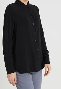 Camicia nera a maniche lunghe con colletto, caratterizzata da una vestibilità comoda e una texture morbida. Indossata con pantaloni grigi chiari.