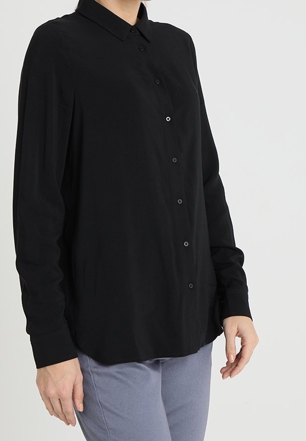 Camicia nera a maniche lunghe con colletto, caratterizzata da una vestibilità comoda e una texture morbida. Indossata con pantaloni grigi chiari.