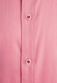 Primo piano di un tessuto rosso testurizzato con due bottoni bianchi cuciti verticalmente su una patta della camicia.