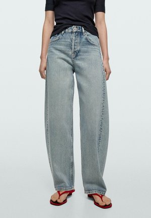 Osoba stojící proti světlému pozadí, oblečená v volných světle modrých džínách s vysokým pasem, černém krátkém tričku a červených sandálech typu thongs.