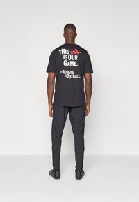 Svart t-shirt med vit och röd text på baksidan som säger "DETTA ÄR VÅRT SPEL. - Adidas Fotboll." Buren med svarta byxor och skor.