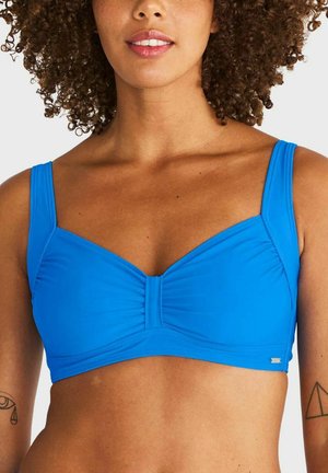 Vrouw met krullend haar draagt een felblauwe bikinitop, toont tatoeages op beide bovenarmen tegen een witte achtergrond.