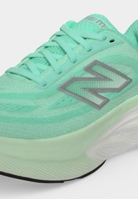 Κοντινή λήψη από ένα αθλητικό παπούτσι New Balance σε μίντ χρώμα με πλέγμα στο πάνω μέρος, επενδυμένο γιακά, κορδόνια και μαξιλαρωμένη λευκή σόλα με μαύρη πέλμα.