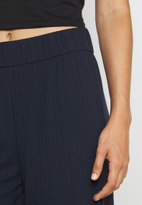 Navy pleated pants featuring a wide waistband and textured fabric, partially shown with a hand resting beside the waistband.