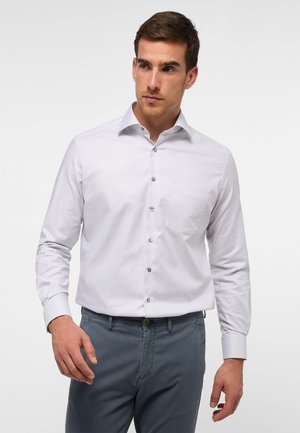 White long-sleeve shirt with a button-up design, single chest pocket, and textured fabric. Grey pants are worn, with a neutral background.