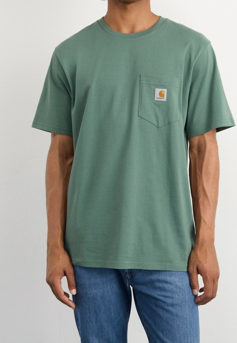 T-shirt verde in cotone con maniche corte, collo a giro e tasca sul petto sinistro con patch logo. Indossato con jeans blu.