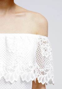 Top bianco senza spalle realizzato in pizzo testurizzato, con bordi scallop e motivi floreali. Tessuto leggero e traspirante.