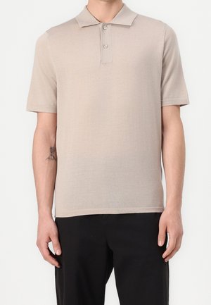 Mand iført en beige kortærmet polotrøje og sorte bukser, stående med armene afslappet ned langs siden mod en ensfarvet baggrund.