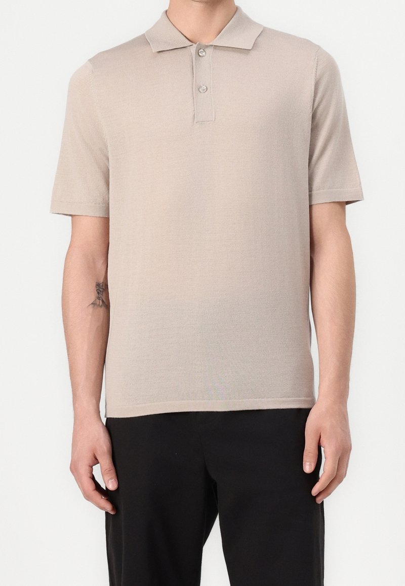 Homme portant un polo beige à manches courtes et un pantalon noir, debout avec les bras détendus le long du corps devant un fond uni.