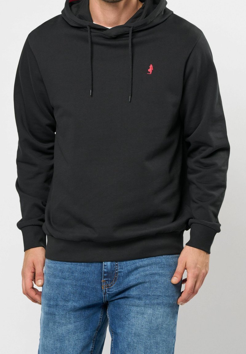 Homme portant un sweat à capuche noir avec un petit logo rouge sur la poitrine, associé à un jean bleu, debout devant un fond uni.